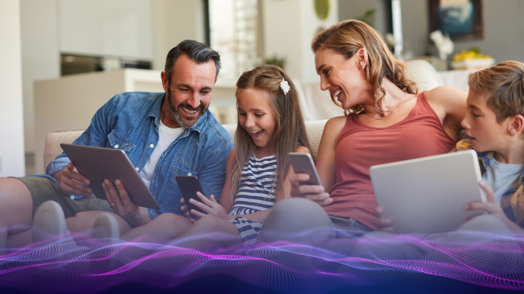 View of a family sitting on a couch and each holding a digital device. They are smiling and laughing at the video displayed on the young girl's device.
