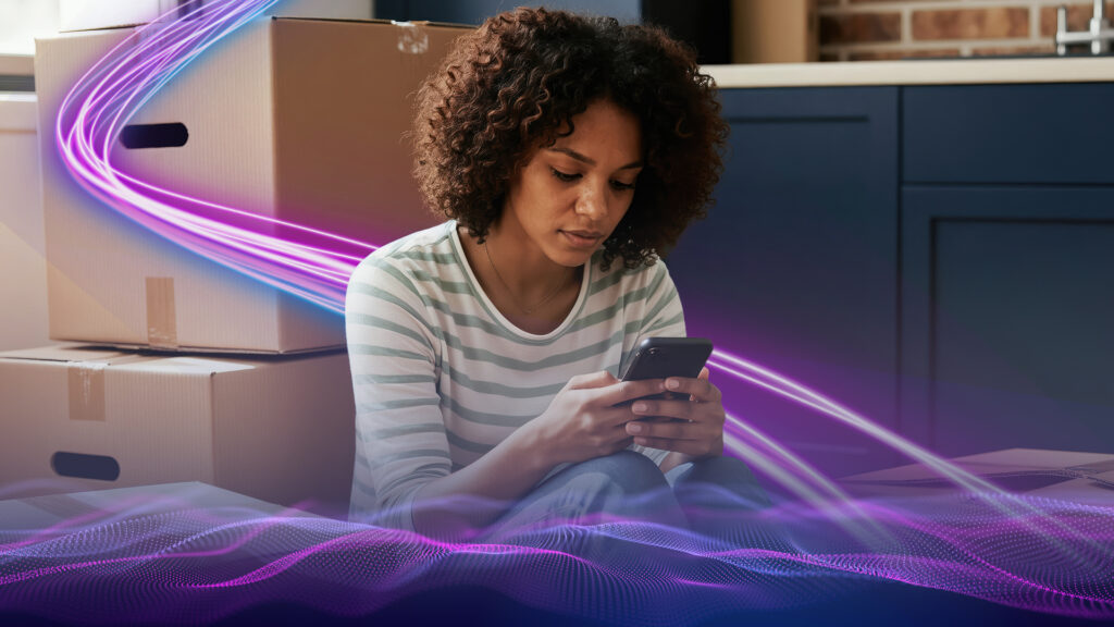 Image of a woman looking at her smart phone with moving boxes in the background.