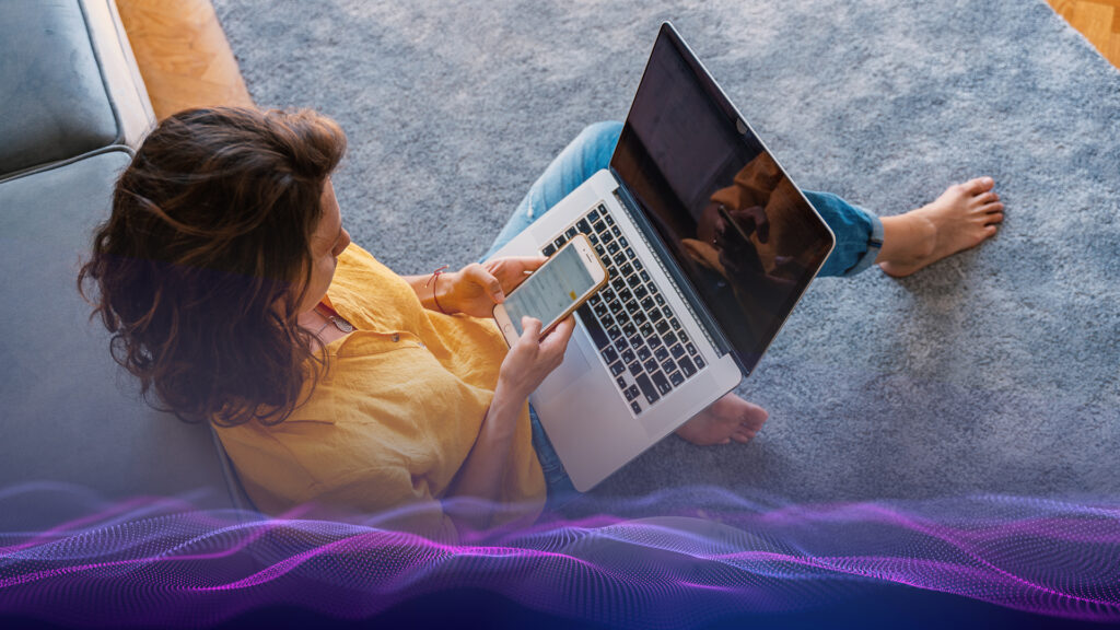 Top down view of a woman sitting on the floor in front of her couch. A laptop is in her lap and she is looking at something on her smart phone.